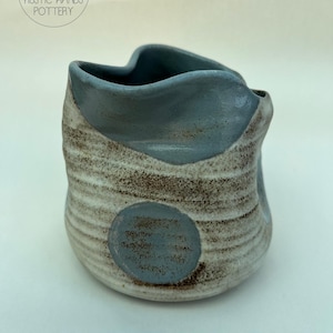 May include: A handmade ceramic vase with a unique, organic shape. The vase features a light blue rim and a circular accent, with a textured, earthy design in shades of brown and cream. The vase is a piece of rustic pottery.