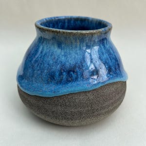 May include: A small, rounded ceramic vase featuring a two-tone design. The upper portion is coated in a glossy, vibrant blue glaze that drips down over the lower section, which consists of a textured, matte dark grey clay surface.