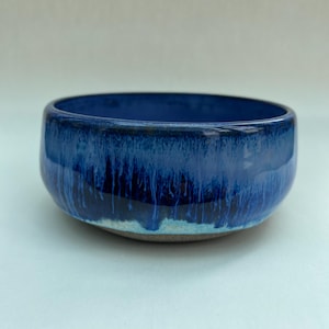 May include: A round ceramic bowl featuring a deep blue glaze with a glossy finish. The exterior displays a vertical drip pattern transitioning from dark navy to a lighter sky blue near the base, which reveals a raw, unglazed brown clay texture.