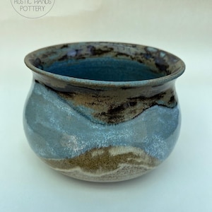 May include: A handmade ceramic bowl with a rounded form and a broad opening. The bowl displays a mix of blue, brown, and white tones, with a shiny finish. The rim is a darker shade of brown. The text "RUSTIC HANDS POTTERY" is visible.