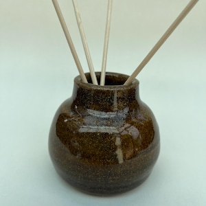May include: A small, round, dark brown ceramic vase with a glossy, speckled finish. The vase has a narrow neck and a wide, bulbous body. Three thin, light-coloured wooden reeds are inserted into the opening, extending upwards from the centre.