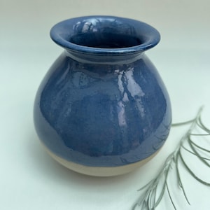 May include: A small, round ceramic vase with a glossy, deep blue glaze covering the upper portion and a smooth, unglazed beige base. The vase features a wide, flared rim and a bulbous body, showcasing a distinct contrast between the blue finish.