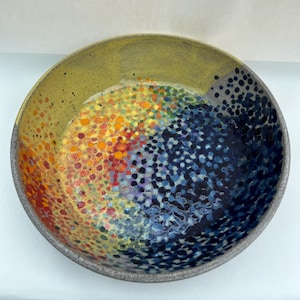 May include: A round ceramic bowl featuring a vibrant interior pattern of multicoloured dots. The design transitions from warm yellow, orange, and red hues on one side to deep blue and black tones on the other, creating a gradient effect across the surface.