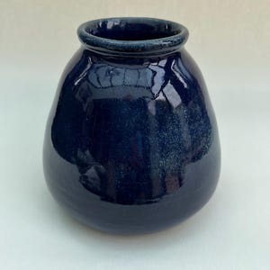May include: A small, bulbous ceramic vase with a deep navy blue glaze. The surface features a glossy finish with subtle, lighter blue speckled variations throughout the body. It has a rounded, thick-rimmed opening and a wide, curved base.