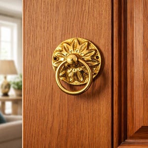 Puede incluir: Un aldaba dorada con un diseño floral y un tirador de anillo está montada en una puerta de madera. La puerta es de color marrón cálido. El fondo está borroso, mostrando un vistazo de una sala de estar.