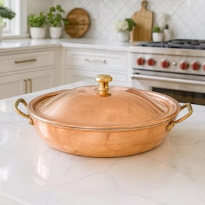 May include: A copper-colored cooking pot with a lid and brass handles sits on a white countertop in a kitchen. The pot has a gold-colored knob on the lid. The kitchen has white cabinets and a stainless steel range hood.
