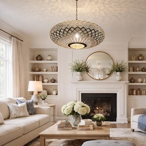 Puede incluir: Un salón luminoso con un sofá beige, un sillón y una mesa de centro de madera. Una gran lámpara de araña adornada cuelga del techo. La habitación cuenta con chimenea, estanterías empotradas y una gran ventana, creando un espacio luminoso y acogedor.