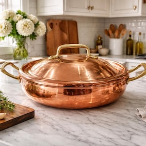 May include: A copper-colored cooking pot with a lid and brass handles sits on a marble countertop. The pot is round and reflective, with a matching lid and a brass handle on top. The kitchen setting includes a cutting board with herbs and a lemon.