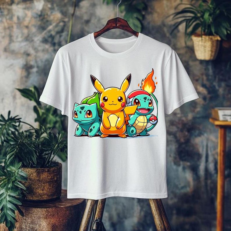 May include: White t-shirt featuring a colorful graphic of three cartoon characters: Pikachu, Bulbasaur, and Squirtle. Pikachu is yellow, Bulbasaur is teal, and Squirtle is blue. The design is centered on the shirt, which is hanging on a wooden hanger.