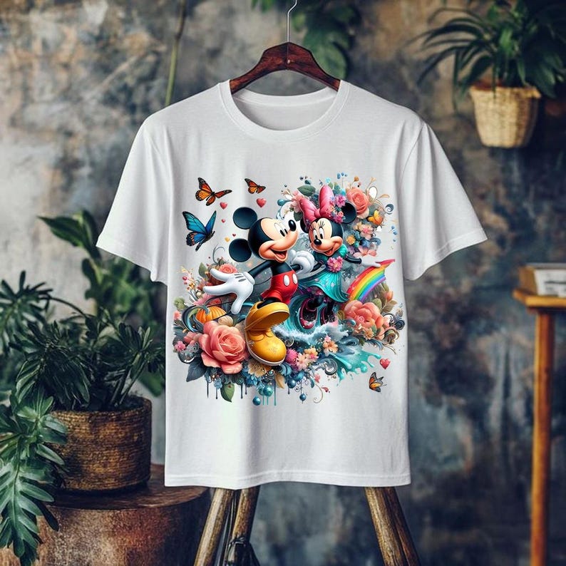 May include: White t-shirt featuring a colorful graphic of Mickey and Minnie Mouse surrounded by flowers, butterflies, and a rainbow. Mickey wears red shorts and yellow shoes. Minnie wears a blue dress and pink bow. The design is vibrant and detailed.
