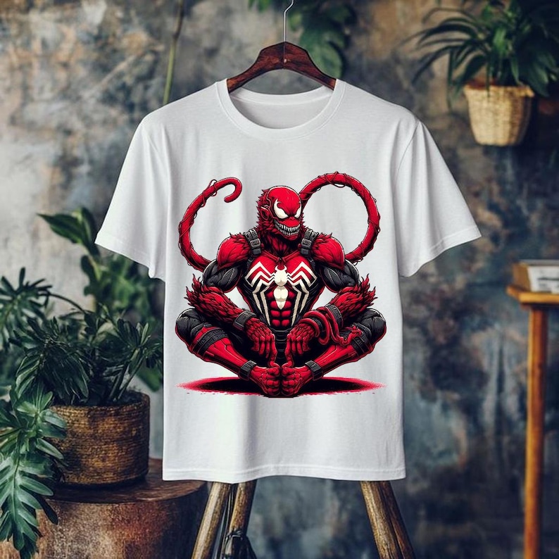 May include: White t-shirt featuring a red and black graphic of a character in a seated position. The character has a red head, black and red body armor, and a white spider symbol on the chest. The design includes long, red tendrils extending from the head.