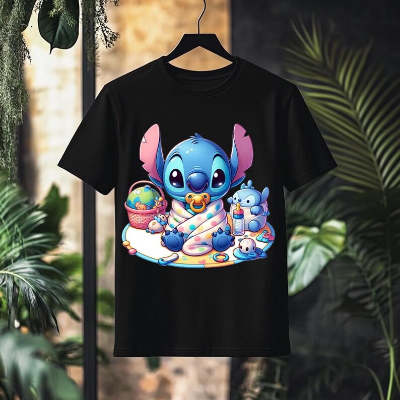 May include: Black t-shirt featuring a cartoon illustration of a blue alien character with large ears, a pacifier, and a blanket. The design includes a basket, a bottle, and other small characters. The shirt is hanging on a black hanger.