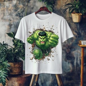 May include: White t-shirt featuring a graphic of the Hulk bursting through a wall. The Hulk is depicted in green with black hair, clenched fists, and an angry expression. The design includes a cracked wall effect.