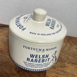 May include: A white ceramic Welsh Rarebit jar with a lid, featuring blue text and a rabbit illustration. The jar is from Fortnum & Mason, made in England. The jar contains 250g of Welsh Rarebit.