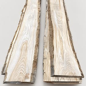 K&ouml;nnte beinhalten: Zwei lange, schmale Holzbohlen mit Naturkante in hellbrauner und cremefarbener Maserung. Die Bretter weisen auf einer Seite eine nat&uuml;rliche, raue Rindenkante auf und haben glatte, flache Oberfl&auml;chen mit komplexen, geschwungenen Maserungen.