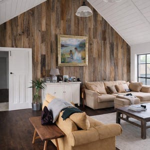 May include: A living room with a feature wall of reclaimed wood panels and a white, vaulted ceiling. The room includes a tan sofa, a wooden coffee table, and a small bench. A framed painting hangs on the wall, and two pendant lights hang from the ceiling.