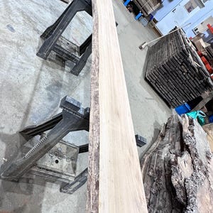 May include: A long, rectangular live edge wood slab with a light tan, smooth surface and a dark, textured natural bark edge. The slab is supported by two black plastic sawhorses in a workshop setting, showcasing the raw grain and wood texture.