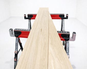 May include: Two long, light-colored wooden planks with visible grain patterns resting horizontally across two red and black metal sawhorses. The planks are positioned parallel to each other, extending from the foreground into the distance.