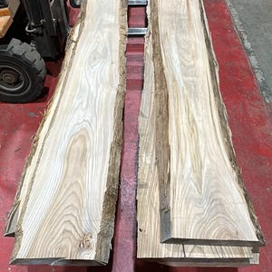 K&ouml;nnte beinhalten: Zwei lange, unbehandelte Holzbohlen mit nat&uuml;rlichen Baumkanten und sichtbarer hellbrauner Holzmaserung. Die Planken liegen parallel zueinander und zeigen eine glatt geschliffene Oberfl&auml;che sowie rohe, mit Rinde bedeckte Seiten.