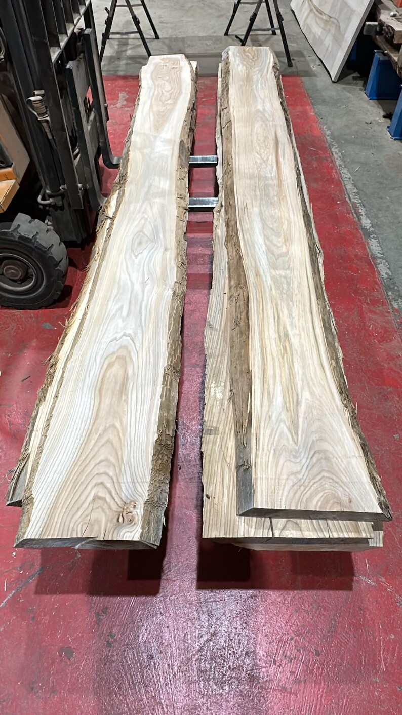 K&ouml;nnte beinhalten: Zwei lange, unbehandelte Holzbohlen mit nat&uuml;rlichen Baumkanten und sichtbarer hellbrauner Holzmaserung. Die Planken liegen parallel zueinander und zeigen eine glatt geschliffene Oberfl&auml;che sowie rohe, mit Rinde bedeckte Seiten.