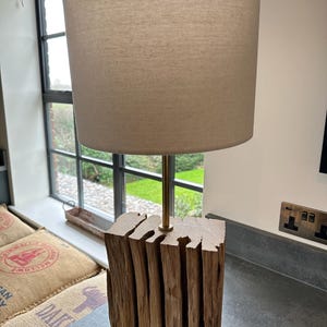 May include: A wooden table lamp with a beige lampshade. The base is made from stacked, roughly cut wood, and the lamp has a brass-coloured stem. The lamp is designed for indoor use and provides ambient lighting.