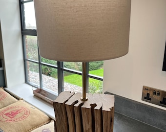 Könnte beinhalten: Rustikale Tischlampe mit beigefarbenem Stoffschirm. Der Sockel besteht aus gestapeltem, grob behauenem Holz, und die Lampe hat einen messingfarbenen Stiel. Die Lampe ist für den Innenbereich konzipiert und bietet Umgebungslicht.