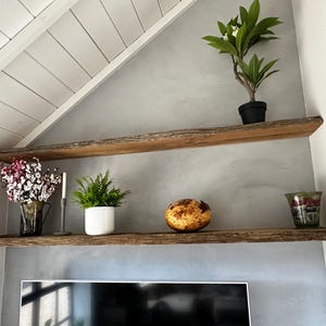 May include: Two horizontal wall-mounted shelves made of natural wood with live edges. The top shelf holds a small potted green plant. The bottom shelf displays a glass vase with pink flowers, a candle, a green plant in a white pot, a round decorative object, and a glass bowl.