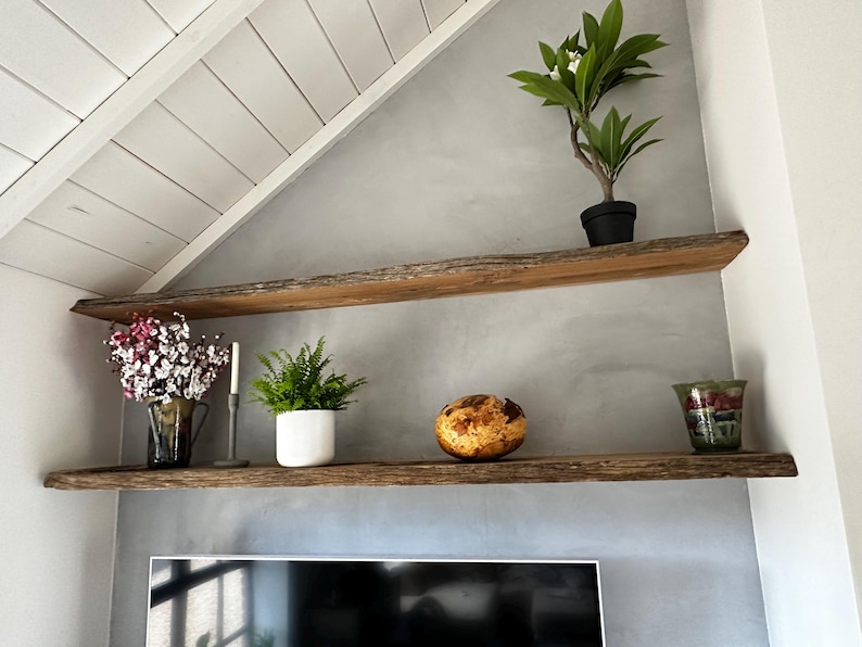 May include: Two horizontal wall-mounted shelves made of natural wood with live edges. The top shelf holds a small potted green plant. The bottom shelf displays a glass vase with pink flowers, a candle, a green plant in a white pot, a round decorative object, and a glass bowl.