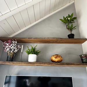 May include: Two natural live-edge wooden shelves mounted on a gray wall. The top shelf holds a small green plant in a black pot. The bottom shelf features a vase with pink flowers, a tall candle, a green plant in a white pot, a round textured object, and a glass.