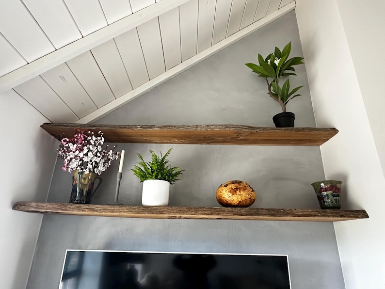 May include: Two natural live-edge wooden shelves mounted on a gray wall. The top shelf holds a small green plant in a black pot. The bottom shelf features a vase with pink flowers, a tall candle, a green plant in a white pot, a round textured object, and a glass.
