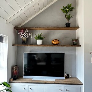 May include: A living room wall featuring two natural edge wooden shelves mounted above a flat screen television. Below the television is a wooden countertop with four white cabinet doors. Various potted plants and decorative items sit on the shelves.