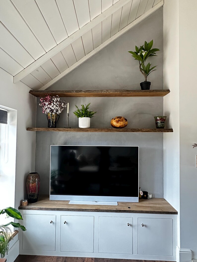 May include: A living room wall featuring two natural edge wooden shelves mounted above a flat screen television. Below the television is a wooden countertop with four white cabinet doors. Various potted plants and decorative items sit on the shelves.