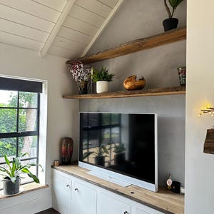 May include: A white media console with a natural wood top and two live-edge wooden shelves mounted on a gray wall. The console features four cabinet doors with silver knobs. A flat-screen television sits on the console, surrounded by potted plants.