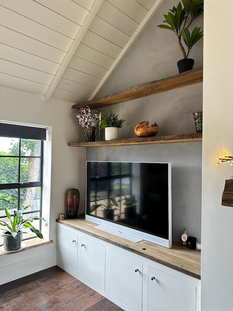 May include: A white media console with a natural wood top and two live-edge wooden shelves mounted on a gray wall. The console features four cabinet doors with silver knobs. A flat-screen television sits on the console, surrounded by potted plants.