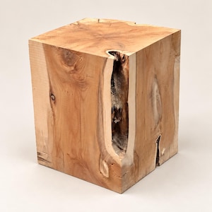 May include: A square wooden stool crafted from a solid block of light brown timber. The piece features a natural wood grain pattern, visible knots, and a deep, vertical fissure along one side that reveals a dark, textured interior.