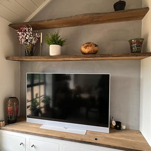 May include: A television set sits on a wooden cabinet with four white doors and silver knobs. Above the television are two natural edge wooden shelves holding a potted plant, a vase of flowers, a fern, a wooden bowl, and a decorative glass vessel.