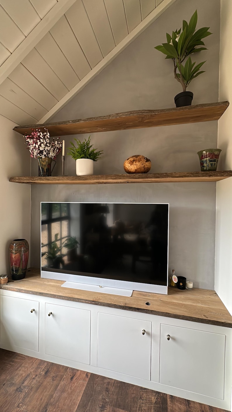 May include: A television set sits on a wooden cabinet with four white doors and silver knobs. Above the television are two natural edge wooden shelves holding a potted plant, a vase of flowers, a fern, a wooden bowl, and a decorative glass vessel.