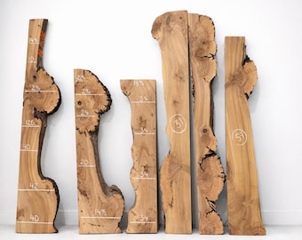 May include: Six vertical live edge wood slabs labeled A, B, C, D, E, and G. Each slab features natural wood grain, knots, and bark edges. White handwritten numbers and horizontal lines are marked on the surfaces to indicate specific measurements.