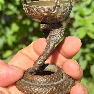 May include: A bronze-toned metal candle holder featuring a coiled snake base with a raised cobra head supporting a small, circular, scalloped-edge bowl at the top. The surface of the metal displays a textured, weathered finish with dark brown patina.