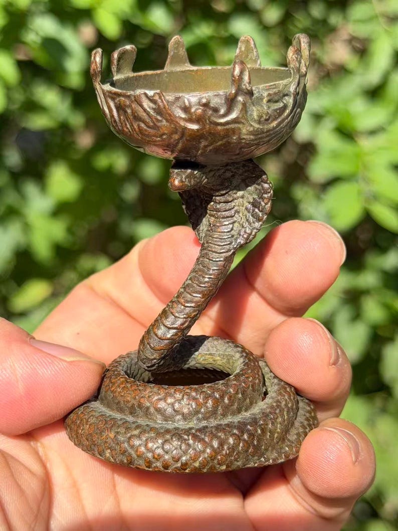 May include: A bronze-toned metal candle holder featuring a coiled snake base with a raised cobra head supporting a small, circular, scalloped-edge bowl at the top. The surface of the metal displays a textured, weathered finish with dark brown patina.
