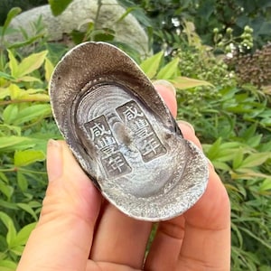 May include: A silver-toned, boat-shaped ingot with a textured, metallic surface. The centre features two rectangular recessed panels, each containing four embossed Chinese characters. The object has a curved, raised rim and a smooth, concave base.