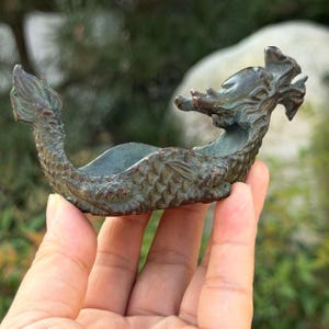 May include: A bronze-toned metal dragon figurine shaped like a small, curved dish. The creature features detailed scales along its body, a raised tail, and a stylized head with prominent horns. The surface has a weathered, dark patina finish.