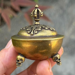 May include: A small, round, antique-style brass incense burner with a lid. The lid features an ornate, raised finial at the top. The base of the bowl is supported by three distinct, sculpted brass skull feet, providing a stable foundation.