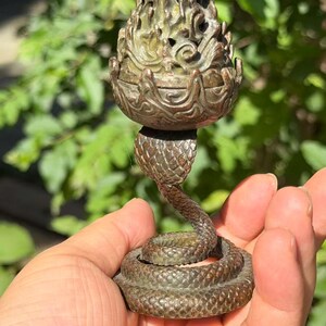 May include: A bronze-toned metal incense burner featuring a coiled snake base with textured scales. The upper portion is a removable lid shaped like a stylized flame or pinecone with intricate cut-out details and a dark, weathered patina finish.