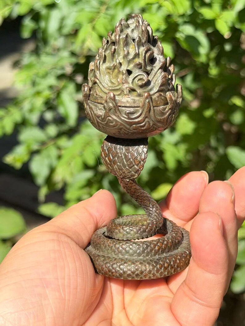 May include: A bronze-toned metal incense burner featuring a coiled snake base with textured scales. The upper portion is a removable lid shaped like a stylized flame or pinecone with intricate cut-out details and a dark, weathered patina finish.