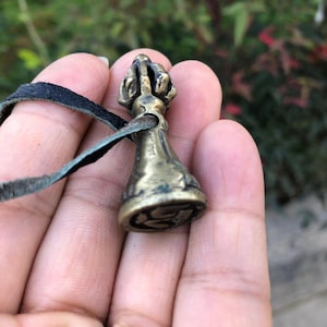 May include: A small, antique brass pendant with a detailed, ornate design. The pendant is bell-shaped with a textured surface and is attached to a black leather cord. The background is blurred, suggesting an outdoor setting.