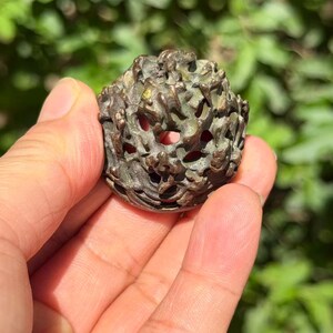 May include: A small, round, dark bronze-toned object with an intricate, openwork, textured surface featuring irregular, raised, branch-like protrusions and small, hollow, circular openings throughout the entire exterior of the piece.