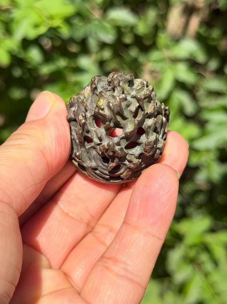 May include: A small, round, dark bronze-toned object with an intricate, openwork, textured surface featuring irregular, raised, branch-like protrusions and small, hollow, circular openings throughout the entire exterior of the piece.
