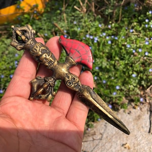 May include: An antique-style dagger in bronze tones, with a red mosaic-style accent. The dagger has detailed carvings and a sharp blade. The handle is intricately designed, and the red accent is at the top. The dagger is held in a hand.