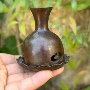 May include: A small, dark bronze-toned metal incense burner featuring a rounded body with a narrow neck and a flared rim. The base of the vessel has a scalloped edge and includes decorative cutouts shaped like stylized clouds or organic forms.